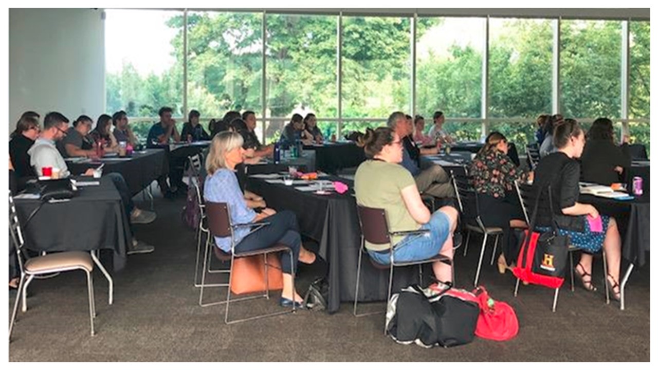 View of a meeting room with large windows looking out onto a forest of trees with numerous tables scattered in a semicircular pattern with numerous people sitting around them. 
