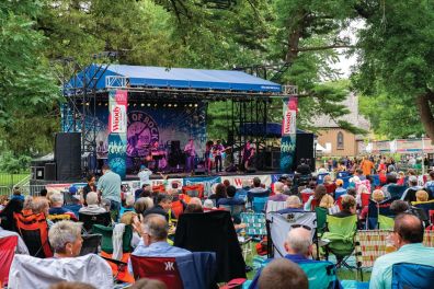 A stage from a distance, with audience members sitting in folding and camp chairs on a lawn.