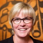 Headshot of a white woman with short blond hair with dark rimmed glasses and wearing dangling earrings. 