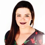 A woman smiles at the camera in front of a white backdrop wearing a floral print dress with her long brown hair swept over her right shoulder. 