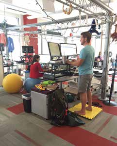 A man stands at a standing desk another walks on a treadmill desk