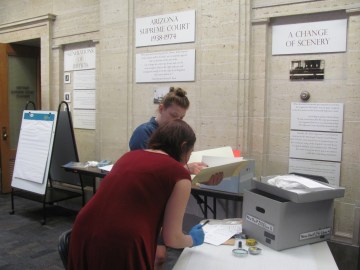 Two women hard at work examining several pieces of archival materials at a table with a grey bankers box on it.