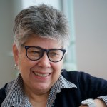 Headshot of Linda Norris, a white woman with short gray/brown hair wearing dark rimmed glasses and a striped shirt under a dark jacket or sweater. 