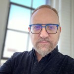 Headshot selfie of Mark Schlemmer, a white man with a full grey beard and mustache wearing blue rimmed glasses and a dark shirt. 