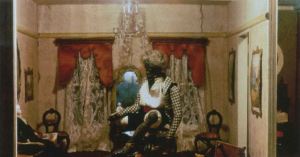 A room in a dollhouse with a black doll in the center and a white doll sitting on furniture in the corner. 