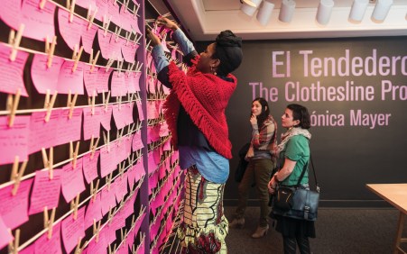 A woman stands on a step stool and clips a pink piece of paper to a wire with hundreds of other pink notes hang. 