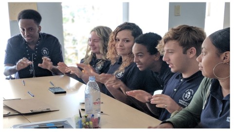 A group of teens sits around a board table their hands prone to smack the table. 