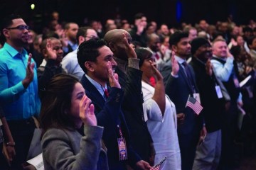 A large group of people stand with their hands raised in a naturalization ceremony. 