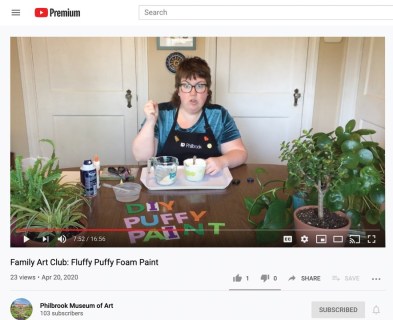 A woman sits behind a desk with a tray of paint in front of her and the words "DIY Puffy Paint" painted on the tabletop in front of her.