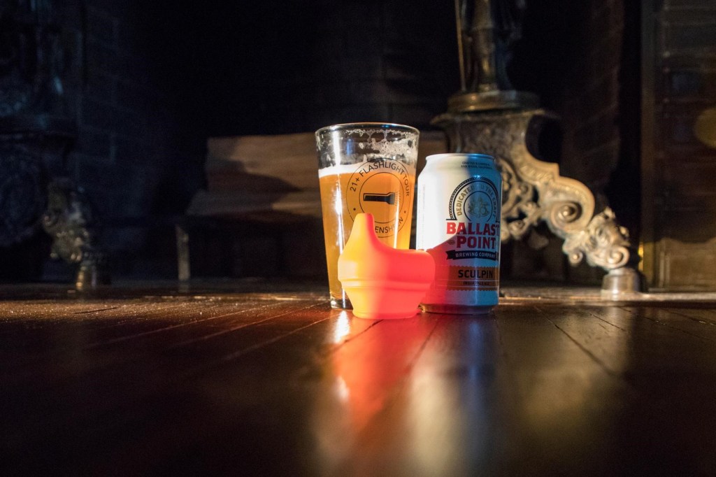 Image taken at floor level of a glass with an amber colored liquid, a can of Ballast Point beer, and a sippy cup lid. 