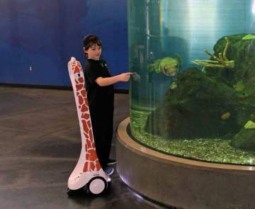 A young man stands next to an aquarium tank with a telepresence robot designed to with giraffe spots