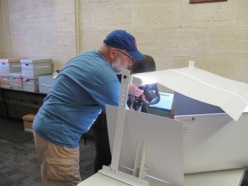 A grey bearded man in a baseball cap holds an object in a lighbox for a female photographer. 