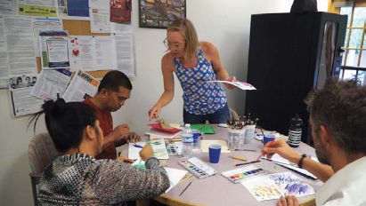 A woman stands at a table surrounded by three other people who are painting on white paper with paint palettes in front of them.