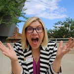 A very excited woman looks as though she is coming toward the camera with her arms close to her body but her hands wide open an expression of awe on her face, mouth open, eyes wide. She has shoulder length blond hair and she is wearing dark rimmed glasses, a black and white striped jacket and a purple top. 