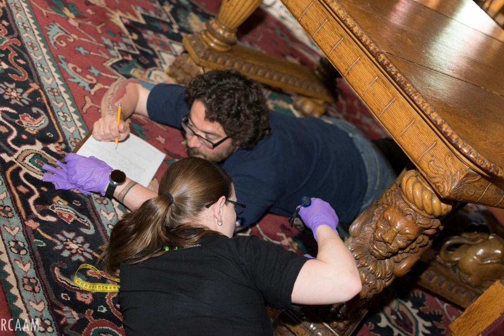 A man lays under an intricately carved table on his stomach filling out a piece of paper and a woman lays near him examining one of the legs. 