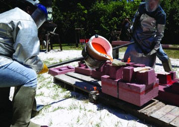 Two men stand next to a large vat of red or pink liquid being poured into a mold.