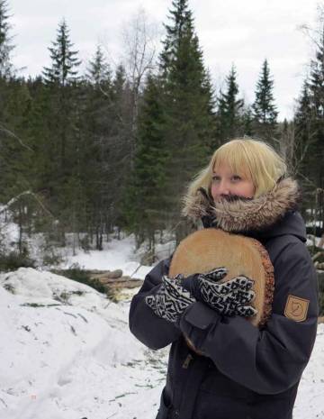 A woman stands in the snow wearing a winter coat and clutching a piece of wood