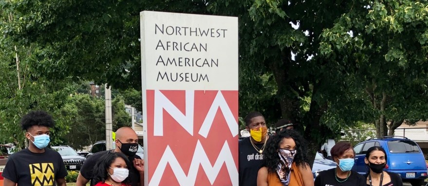 A group of people wearing masks and t-shirts with the museum's logo in front of a sign for the museum in a parking lot.