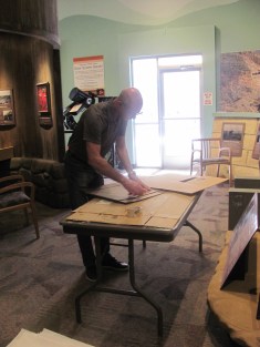 A bald man stands over a folding table working with the back of a picture frame. 