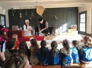 A group of school children sit in front of a demonstration of historic cooking methods.