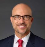 Headshot photo of a man looking at the camera smiling wearing glasses a suit with a red tie