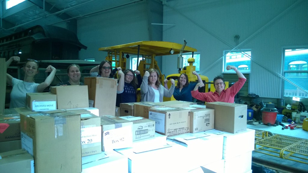 A group of volunteers stand behind a large group of cardboard boxes with their arms raised a la Rosie the Riveter.