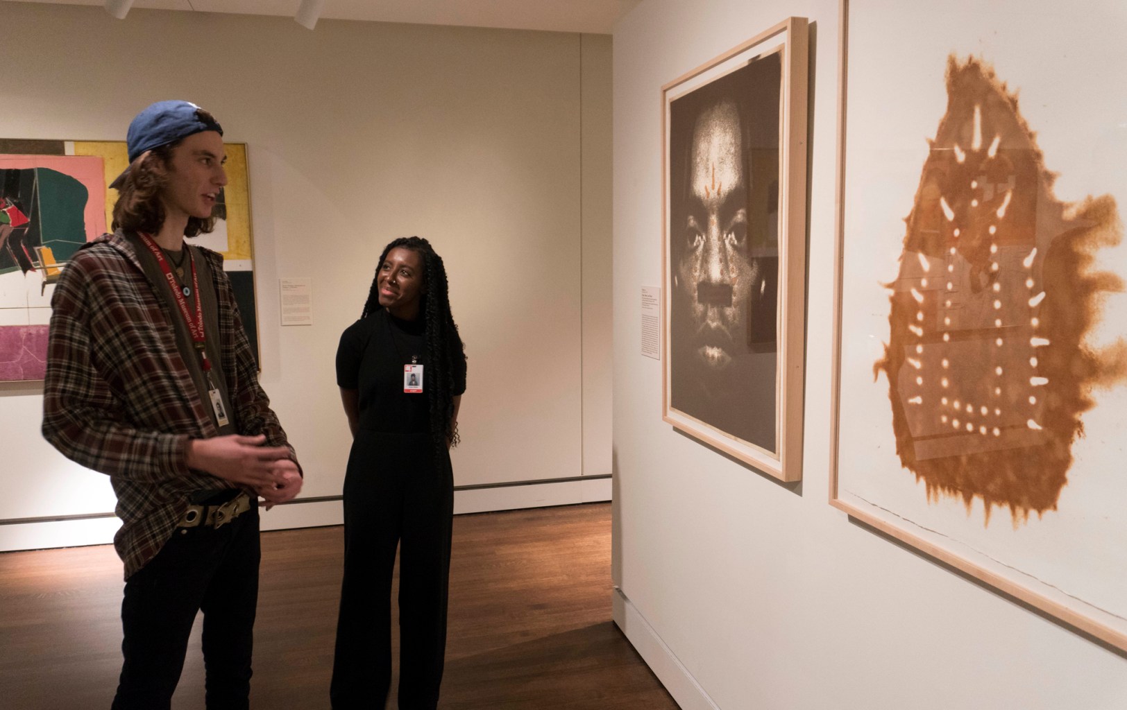 The fellow Asmaa Walton is seen in a gallery space, looking at two hanging works with a young man who also wears a museum staff badge. One of the works is a photograph of a black man's face, and the other is the brown imprint of a clothing iron against a white background.
