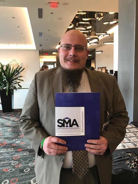 A man in professional dress stands in the lobby of a meeting space holding a folder with the Small Museum Association logo on it.