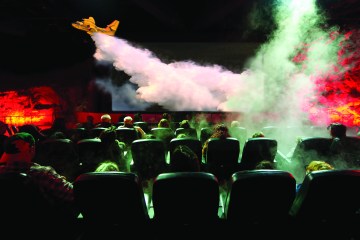 A audience in a theater watches as an airplane releases smoke suppressant over a fire.