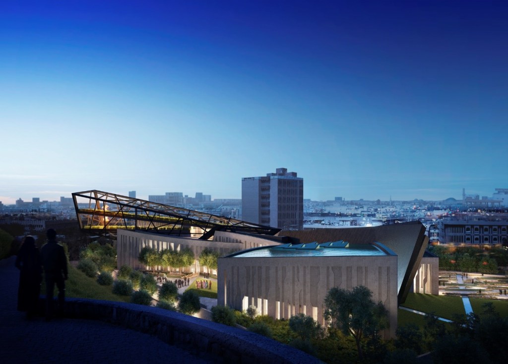 An exterior view of the museum in evening light. The building is made up of slanting, diagonal lines and has large windows around the edifice. The building looks out over a sprawling cityscape.