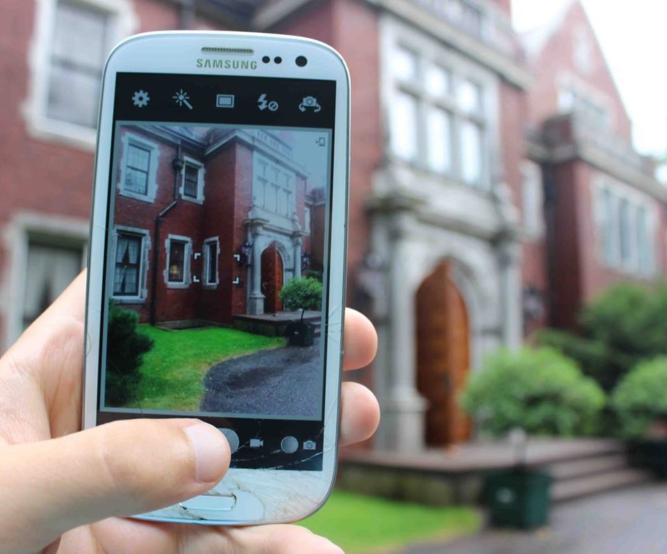 Image of someone holding up their cell phone to take a picture of the outside of Glensheen mansion. You can see the house blurry in the background and clear on the phone screen in the image. 