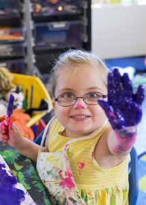 A little girl holds her hand up. It is covered in purple finger paint