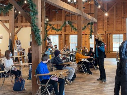 A group of students sit at desks while an instructor teaches before them.