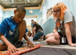 A young black boy and an orange haired white woman sit on the floor working on a yarn loom.