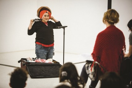 A person kneels in front of an open suitcase in the white space with a microphone in front of them. 