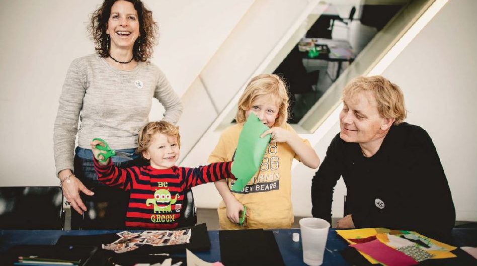 A young family looks towards the camera. Mom is smiling broadly with shoulder length curly hair,. Two young boys interact with paper materials in front of them, each holds a pair of scissors one has a piece of paper in front of his face. The father looks on from a seated position wearing a black long sleeve shirt, he has medium length blond hair.