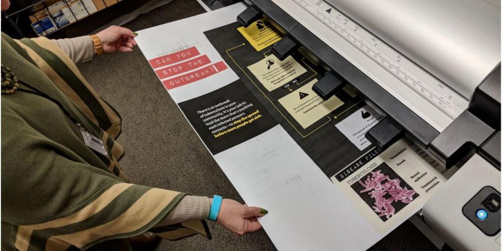 Person in green pulling large poster paper out of printer