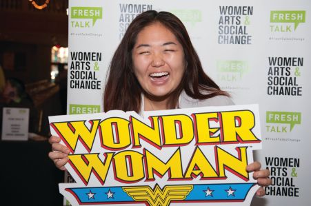 A woman stands with a Wonder Woman sign laughing at someone off camera. 
