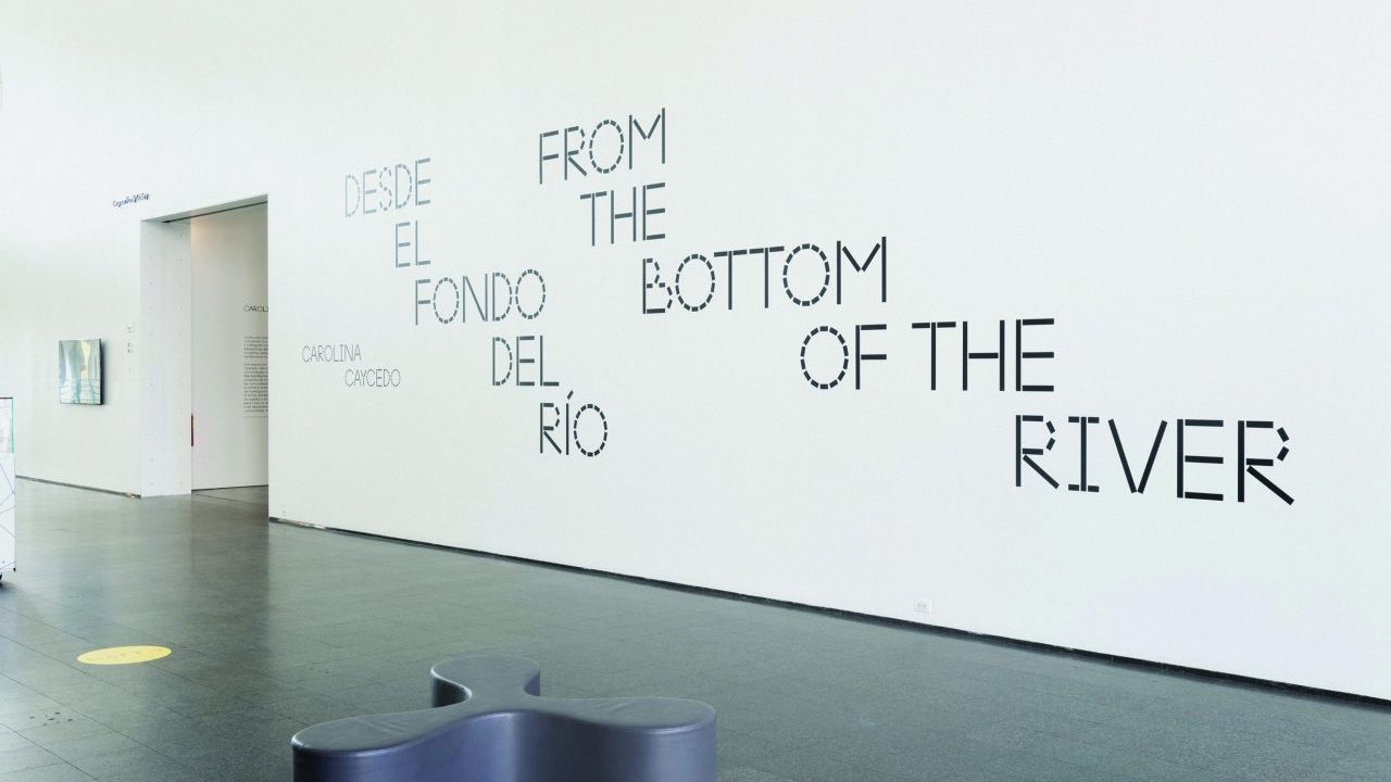 Interior of the Museum of Contemporary Art Chicago with the exhibition title 'Carolina Caycedo: From the Bottom of the River' displayed in large letters on a white wall, in both Spanish and English. A gray, wavy bench is in the foreground.