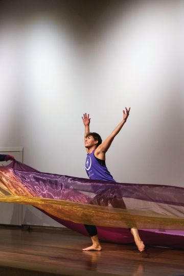A person stands behind a colorful sheer piece of fabric that is blowing in the wind with their arms outstretched as though taking flight.