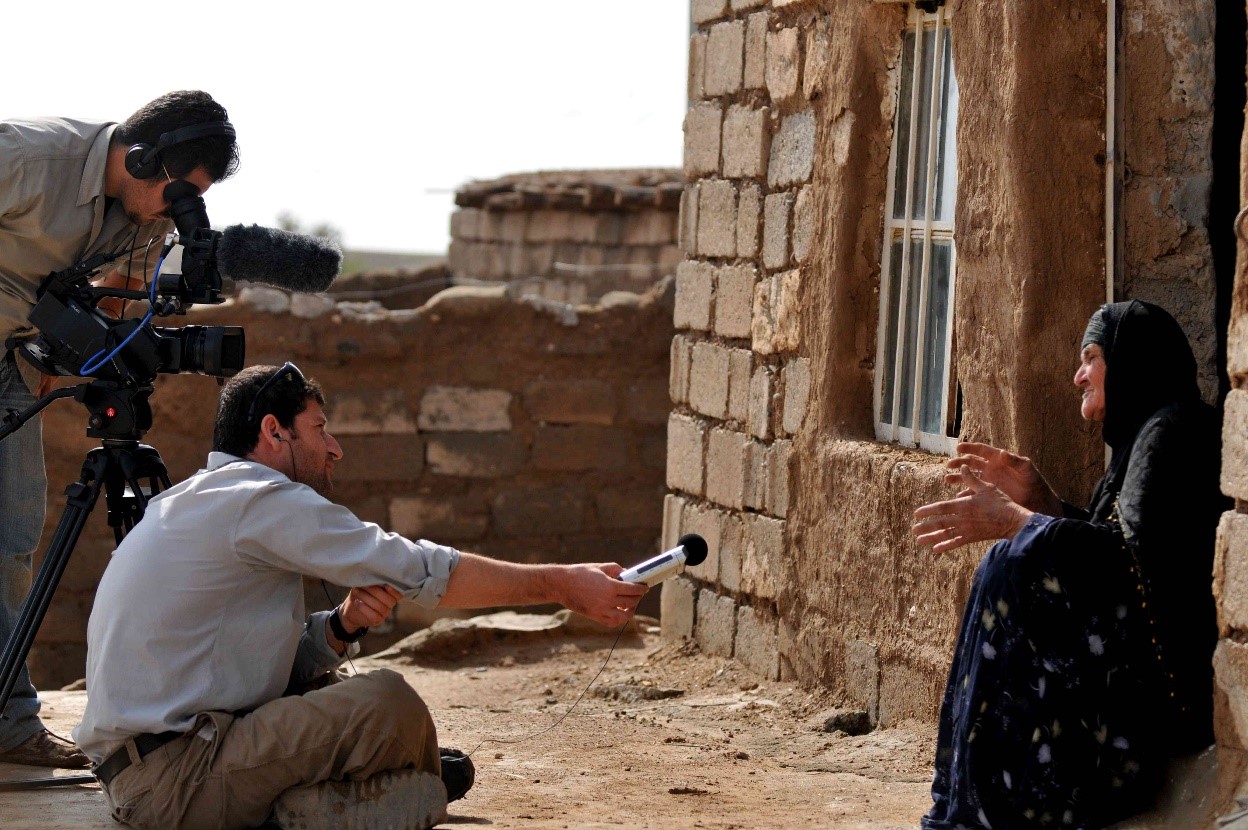 A man holds a microphone toward a woman while a camera operator films her next to him. The woman is wearing traditional Kurdish clothing and sitting in front of a traditional house. The men are in Western clothing.
