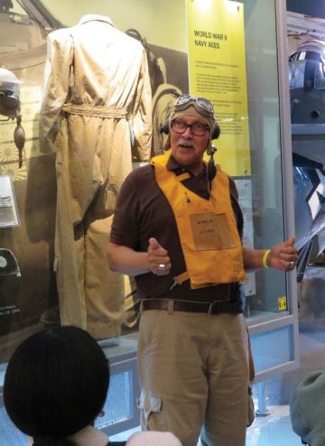 A grey haired man wearing a deflated flotation device stands in front of a museum case and appears to be giving a tour.