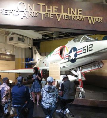 A female tour guide stands in front of a fighter jet with a group of uniformed people in front of her.