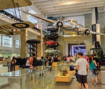 Interior shot of the inside of a museum exhibit with several visitors mulling around a silver airplane hangs from the ceiling and a stack of VW bugs is visible in the corner. 