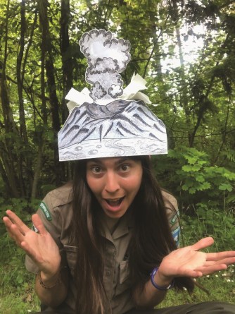 A woman wears a volcano erupting hat on her head with a "wow" expression on her face and her hands outstretched.