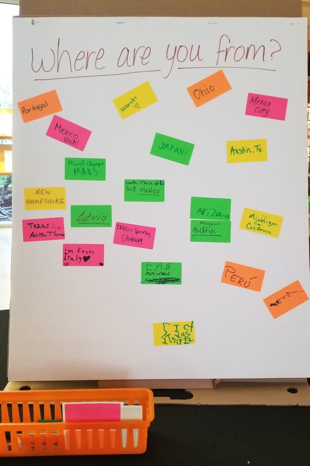 A large sheet of paper with "Where are you from?" written at the top, with colorful sticky notes below that say answers like "Portugal," 'Nebraska," "Ohio," "Mexico City," "Mexico / VIVA!," "JAPAN!," and "Austin, Tx."