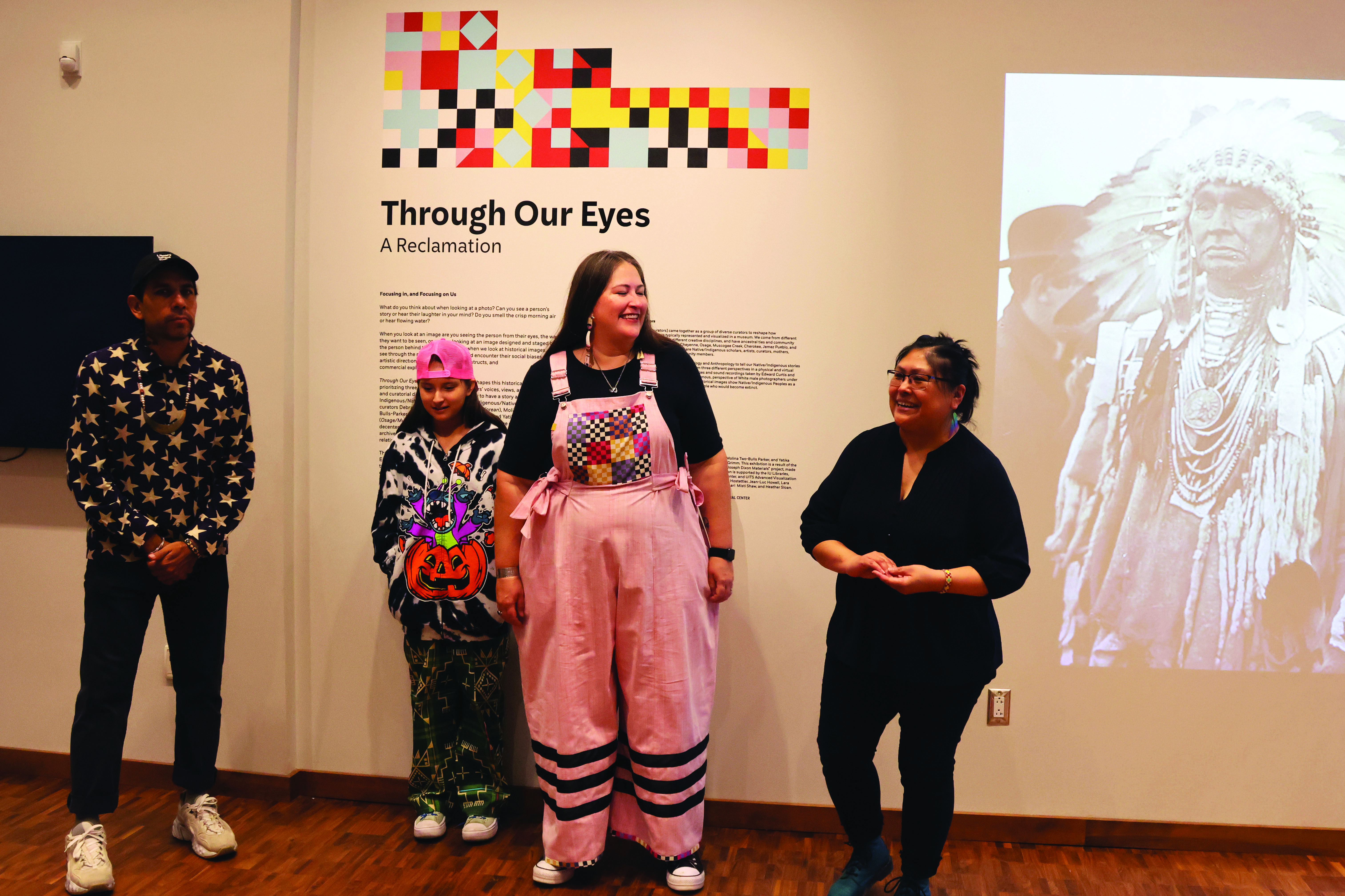A group of people standing in front of a gallery wall with the title "Through Our Eyes: A Reclamation"