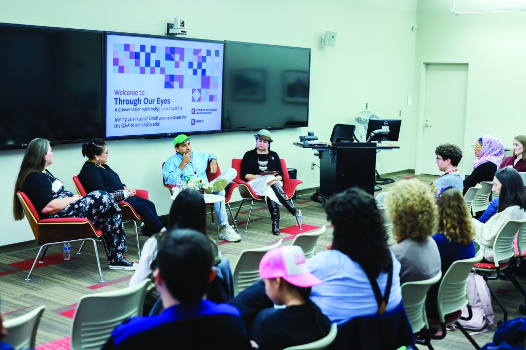 A group of people seated in a lecture room in front of a screen reading "Welcome to Through Our Eyes: A Conversation with Indigenous Curators."
