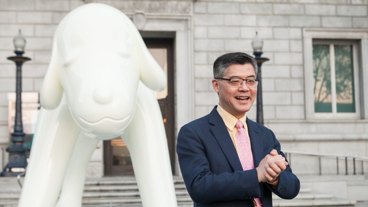A person smiling with clasped hands in front of a stylized statue of a dog