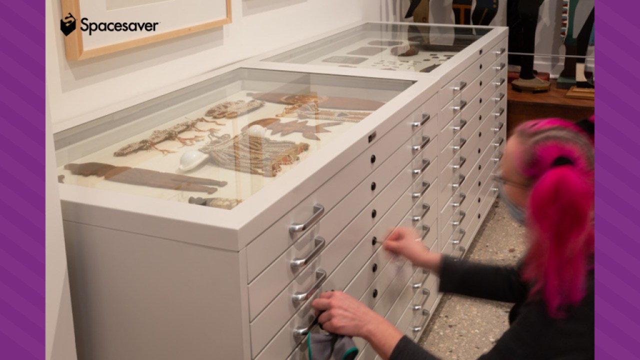 A person unlocking a flat file storage shelving unit with a key, with objects visible in glass window on top